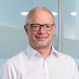Ein Mann mit kurzen grauen Haaren und Brille lächelt. Er trägt ein weißes Hemd. Im Hintergrund ist eine verschwommene Innenkulisse mit Glaswänden und kreisförmigen Lichtern zu sehen.